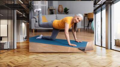 Online home fitness. Senior woman exercising to sports video on laptop in living room, looking at camera and smiling Wall mural