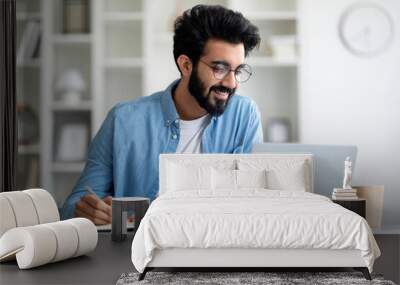 Online Lesson. Smiling Young Indian Man Using Laptop And Taking Notes Wall mural