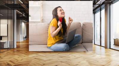 Online shopping. Emotional girl with bank card and laptop computer buying something via internet at home Wall mural