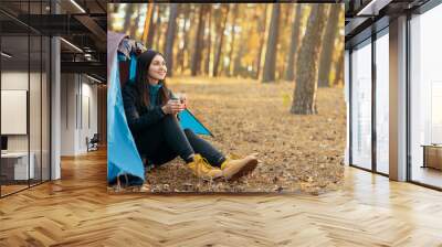 Peaceful young woman sitting in tent, drinking tea and looking around in forest, copy space Wall mural