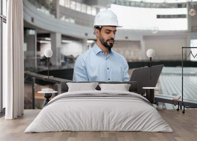 Pensive busy millennial arab guy engineer in safety helmet with beard works with computer controls factory Wall mural