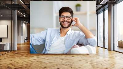 Phone conversation concept. Cheerful arab man talking on smartphone resting on sofa at home interior Wall mural