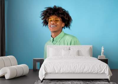 Portrait of happy african american teen guy in mint shirt and yellow sunglasses posing on blue studio background Wall mural