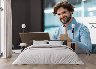 Portrait of happy businessman sitting at desk, using laptop computer and showing thumb up gesture, working at office Wall mural