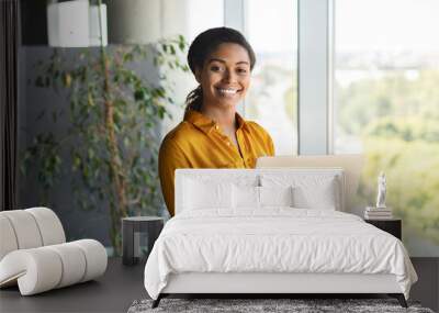 Portrait of happy businesswoman using laptop in office interior, standing near window and smiling at camera Wall mural