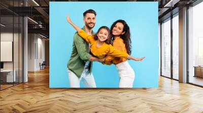 Portrait of happy european parents fooling with their daughter, holding child on hands and girl pretending flying Wall mural