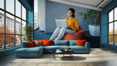 Positive black female manager working on laptop computer in office, sitting on pouf beanbag chair, free space Wall mural