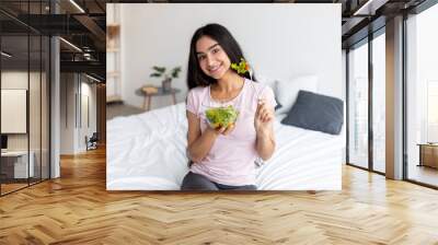 Positive Indian woman sitting on bed and eating delicious vegetable salad at home. Healthy diet for weight loss concept Wall mural