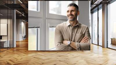 Positive middle aged businessman standing near window in company office, looking away and smiling, copy space Wall mural