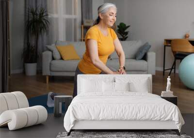 Positive sporty senior woman keeping fit at home, doing lunges, exercising on yoga mat in living room and smiling Wall mural