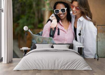 Positive stylish young millennial diverse ladies tourists in hat and sunglasses hugging and looking at map Wall mural