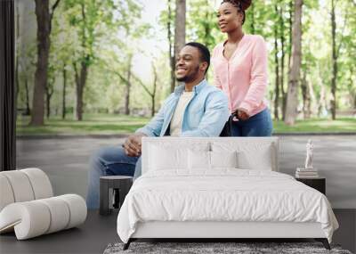 Positive young African American couple with beautiful woman and millennial guy in wheelchair going for walk at park Wall mural