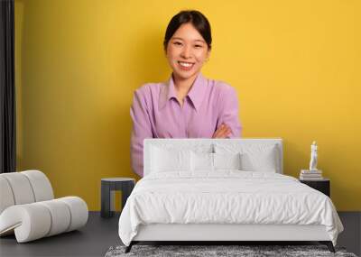 Positive young Asian woman standing with crossed arms and looking to camera over yellow studio background Wall mural