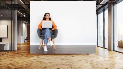 Positive young eastern woman sitting in armchair, using laptop Wall mural