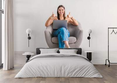 Positive young woman with laptop working online, sitting in armchair and showing thumbs up with both hands Wall mural