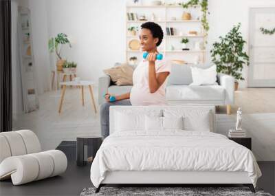 Pregnant black woman sitting on fitness ball, working out with dumbbells in her home gym Wall mural