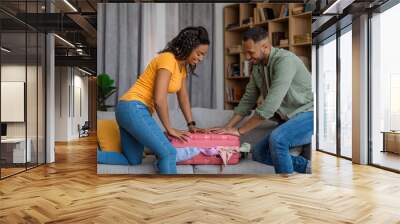 Preparation for vacation. Excited black couple packing and trying to close suitcase on sofa, preparing for summer trip Wall mural