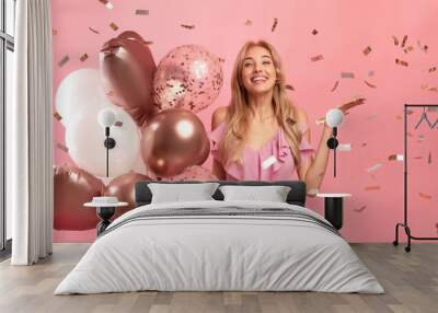 Pretty young woman holding balloons at celebration party, posing over pink studio background with falling confetti Wall mural