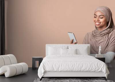 Relaxed black muslim woman in headscarf using smartphone, texting or browsing new app, standing over yellow background with free space Wall mural