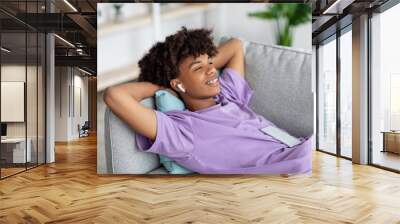 Relaxed black teenager in earphones listening to music or audio book, lying on couch at home Wall mural