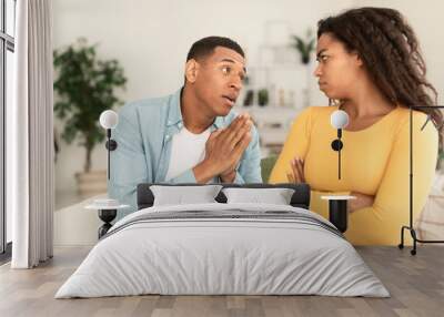 Sad young african american man asking for forgiveness from angry offended female in living room interior Wall mural