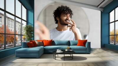 satisfied indian man smelling perfume, doing morning beauty routine, enjoying pleasant odor, standin Wall mural