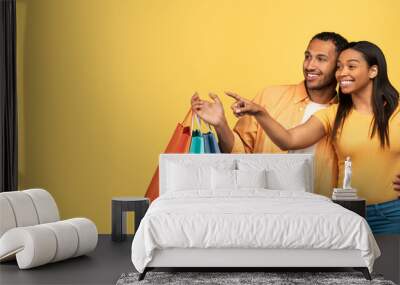 Seasonal sales. Happy young African American couple holding bright shopping bags, pointing aside at empty space Wall mural