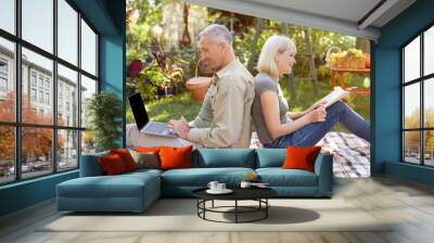 Senior spouses resting in garden, man using laptop computer, woman reading book, sitting back to back, side view Wall mural