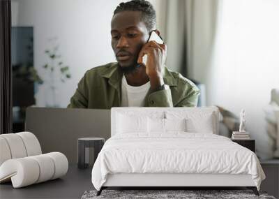 Serious african american male freelancer talking on cellphone and looking at laptop computer, working online from home Wall mural