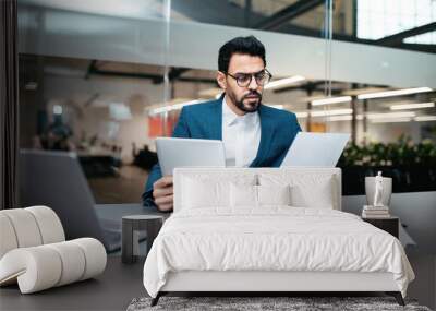 Serious busy handsome young muslim guy with beard in glasses, suit sits at table with computer and tablet Wall mural