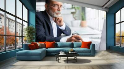 Serious confident pensive european senior businessman in suit looks at laptop with pen at table in office Wall mural