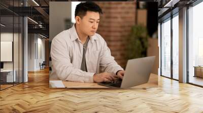 Serious middle aged asian man using laptop computer, typing on keyboard while working online, sitting at home office Wall mural