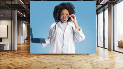 Shocked young african american curly lady doctor therapist in white coat with laptop takes off glasses Wall mural