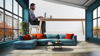 Side view of millennial black businessman using laptop while sitting at desk against white studio wall, copy space Wall mural