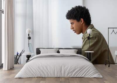 Side view of millennial black student sitting at desk with laptop, making notes in copybook, learning remotely from home Wall mural