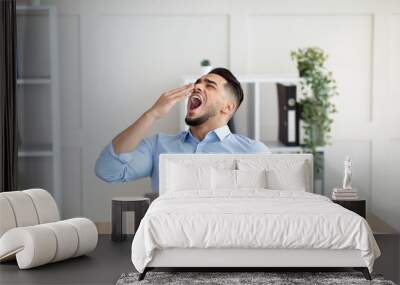 Sleepy young Arab guy yawning, tired of working with laptop at modern office Wall mural