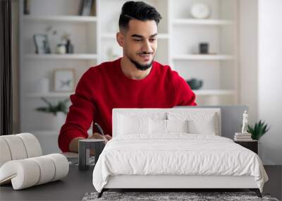 Smiling calm confident attractive millennial arab guy with beard in red clothes makes notes, works on laptop Wall mural