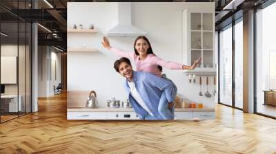 Smiling handsome millennial european husband in casual carries wife on his back and have fun in scandinavian kitchen Wall mural