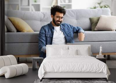 Smiling Indian man using laptop while sitting cross-legged on floor at home Wall mural