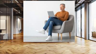 Smiling mature man using laptop sitting on armchair in living room. Happy confident senior male adult resting at home working on pc, typing on keyboard isolated on white studio wall, full body length Wall mural