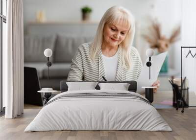 Smiling mature woman writing and holding document at home Wall mural