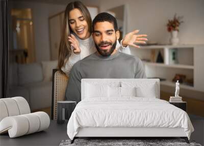 Smiling millennial european female opens eyes with hands to arab husband, gives box with present Wall mural