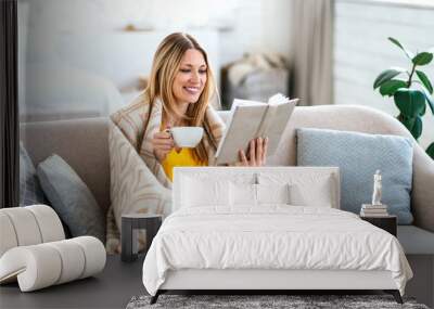 Smiling pretty young european blonde lady wrapped in plaid drinks favorite hot drink and reads book on sofa Wall mural