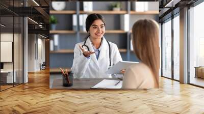 Smiling young attractive indian lady medical worker in white coat gestures and consultation patient at workplace Wall mural