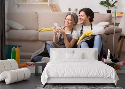 Smiling young couple relaxing with smartphone after cleaning apartment Wall mural