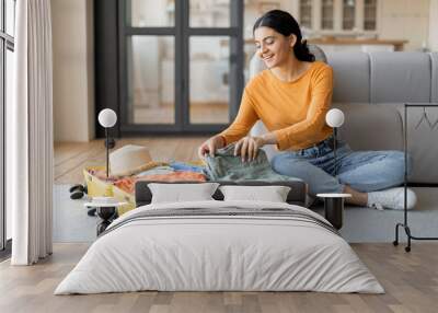 Smiling young indian woman happily packing suitcase at home Wall mural