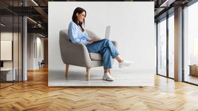 Smiling young woman in casual wear working online, sitting in armchair and using laptop against white wall Wall mural