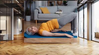 Sporty aged woman doing half bridge yoga pose, strengthening her abs muscles, training in living room, copy space Wall mural