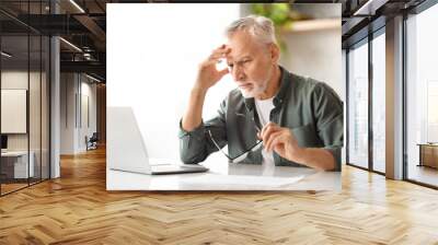 Stressed senior man using laptop while sitting at desk in kitchen Wall mural