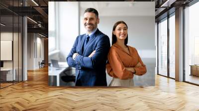 Successful businessman and businesswoman standing back to back with arms crossed and smiling at camera, office interior Wall mural